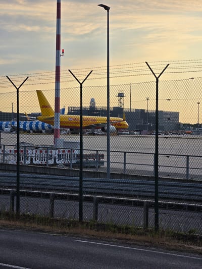 The image captures a view of Frankfurt Airport (FRA) in Kelsterbach, Germany, seen through a chain-link fence with barbed wire at the top. In the foreground, a dark asphalt road with a white line runs along the bottom. Behind the road and fence, a DHL cargo plane, predominantly yellow with red accents, is parked on the tarmac. To the left of the DHL plane, another aircraft with a distinctive blue and white striped livery is visible. Several structures, including airport buildings, lighting towers, and possibly a control tower, are present in the background. A red and white striped pole with a red light is positioned in the mid-ground. The sky is overcast with soft, warm lighting, suggesting either dawn or dusk. The overall scene depicts a quiet, operational area of the airport.