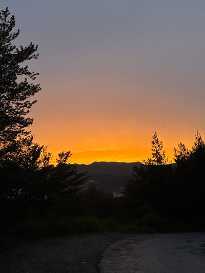 The image depicts a serene natural landscape at dusk in Strazha, Bulgaria. Silhouetted trees frame the foreground, with a dirt road leading into the scene. Beyond the trees, rolling hills are visible against a gradient sky that transitions from a deep blue at the top to a vibrant orange and yellow near the horizon, indicating the setting sun. The overall atmosphere is tranquil and picturesque.