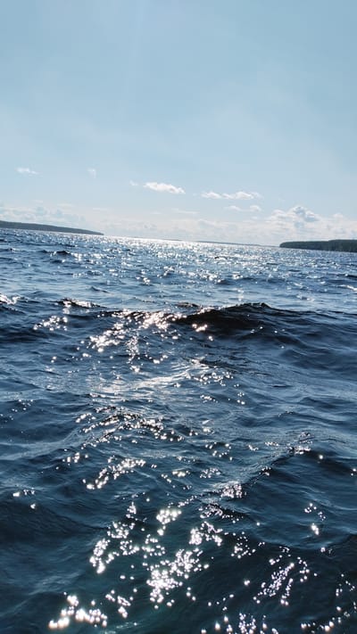 The image depicts a sun-dappled body of water, likely a lake or calm sea, under a bright, partly cloudy sky. The water is a deep blue with numerous small, sparkling reflections of sunlight on its surface, indicating a sunny day with light waves. In the distance, a landmass covered in dense, dark green trees forms a horizon line. The scene appears to be taken from a low vantage point, possibly from a boat or the shore. The location is identified as Nadvoitsy, Russia. There are no people, animals, or man-made objects visible in the frame.