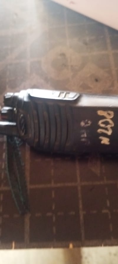 The image shows a close-up of a dark-colored object with ribbed detailing and some markings on it. The object appears to be lying on a textured surface with faint grid lines. The markings on the object include what looks like the letters "PCTA" in a light color and some smaller, less distinct characters. The background is blurred and appears to be a light-colored surface with some shadowing. The image quality is low, with significant blur and low resolution, making precise identification difficult. The location is not discernible from the image itself.