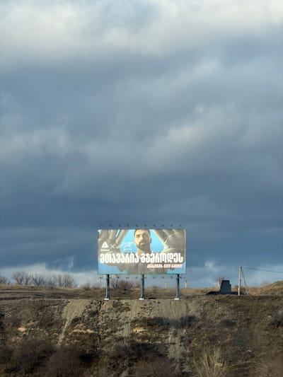 This image depicts a wide outdoor scene in Akhalsopeli, Georgia, under an overcast sky. The upper two-thirds of the frame are filled with heavy, grey-blue clouds. A large rectangular billboard, supported by four dark poles, stands prominently on an elevated, barren, and rocky hillside in the lower third of the image.
The billboard displays an advertisement featuring a man with a dark beard and dark hair, looking directly forward. His head is centered between two large hands, palms open and facing forward, positioned on either side of his head. The background of the advertisement behind the man is a light blue. White Georgian script is visible on the lower left portion of the billboard, accompanied by a white geometric logo and a stylized signature.
The hillside itself is characterized by dry, light brown earth and sparse, dormant vegetation, including bare trees and shrubs, particularly visible along a distant horizon line on the left. The ground slopes downwards sharply from the billboard's base. To the far right, on the same elevated plane as the billboard, a utility pole with wires and a small, dark, box-like structure are present. No other people, vehicles, or specific activities are visible in the scene beyond the billboard's content.