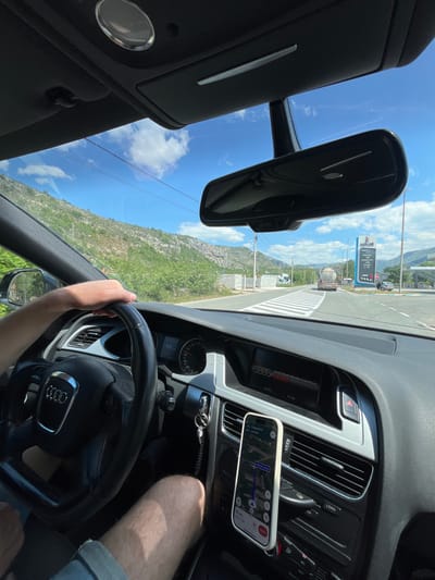 A first-person shot from the driver's seat of an Audi car. The driver's left hand is on the steering wheel. The dashboard contains the Audi Multi Media Interface (MMI) display showing "21.5°C" and "11:38," next to it is a cell phone with a navigation app displaying a turn-by-turn route. Through the windshield, a rural road bordered by green shrubs and mountains is visible. A "klikoil" sign stands roadside. The scene is located in Donje Mrke, Montenegro.