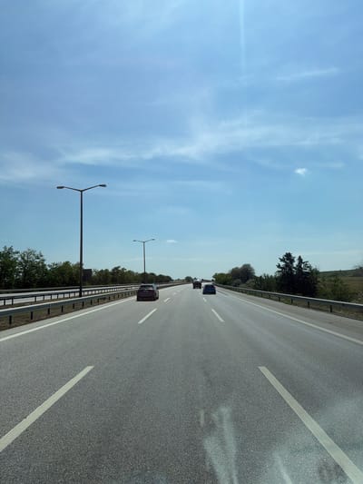 The image displays a multi-lane asphalt highway stretching into the distance under a bright, clear blue sky with sparse, wispy white clouds. The perspective suggests the photo was taken from a moving vehicle.

On the paved road, white dashed lines separate the lanes. Two dark-colored cars are visible ahead, traveling in the same direction. A dark sedan is closer in the right lane, and another car is further ahead in the left lane.

Along the right side of the highway, a metal guardrail runs parallel to the road. Beyond the guardrail, there's a strip of lush green trees and vegetation, with some taller trees and distant hills visible in the background. Two distinct, dark-colored streetlights with Y-shaped tops are positioned along the roadside, extending into the frame from the right.

The scene depicts daytime travel, likely morning or afternoon, given the strong illumination and the slight lens flare visible in the upper left of the sky, indicating the sun's position. The weather appears fair and sunny. The activity taking place is driving on a highway. The location context provided is Edirne Merkez, Turkey. There is no visible text in the image.