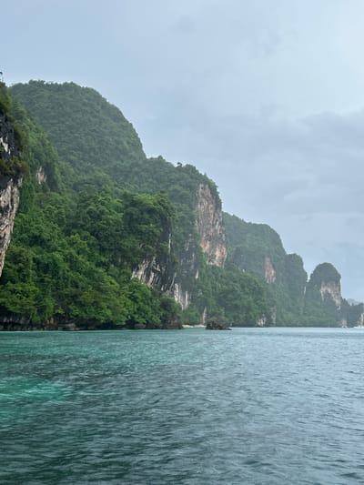The image depicts a scenic view of limestone karsts covered in lush green vegetation, rising dramatically from the turquoise waters of the sea. The karsts are characterized by their steep, rugged cliffs and dense foliage. In the foreground, the water exhibits a gradient of colors, shifting from a clear, aquamarine hue near the shore to a deeper, darker blue further out. The sky is overcast with a pale, grayish hue, suggesting a cloudy or potentially rainy day. The scene conveys a sense of natural beauty and tranquility. The specific location is identified as Haad Hin Khom, Thailand.