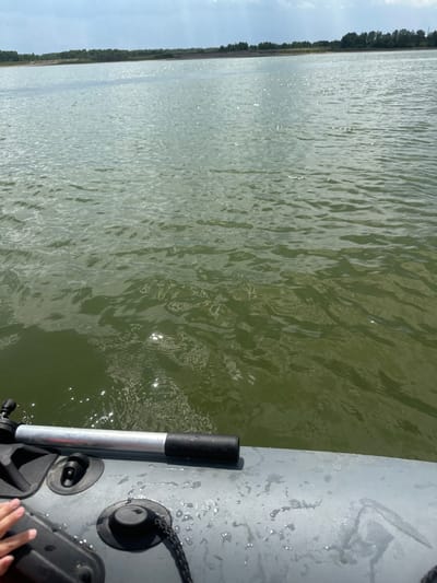 The image shows a view from a small inflatable boat on a body of water. The water is a murky green color and has ripples on its surface, reflecting sunlight. In the foreground, the grey, textured surface of the boat is visible, along with a metal oar handle and black plastic fittings. A person's hand is resting on the boat's surface on the left side. In the distance, across the water, a tree line and a hazy sky are visible. The location is identified as the city of Pravda, Russia.