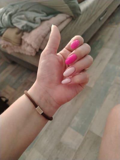 The image features a close-up view of a person's left hand, with a thumb extended upwards and the fingers curled. The hand has neatly manicured nails, with the index finger nail painted a bright pink, and the remaining visible nails painted a pale pink. A brown cord bracelet with a rectangular gold-colored clasp is worn around the wrist. The background shows a blurred scene with what appears to be a pile of blankets or clothing in soft pastel colors, including pink and a muted green or gray. The surface beneath the hand and bracelet is a wooden floor with a planked pattern and a muted color scheme, possibly indicating an indoor setting. The lighting suggests a softly lit environment, possibly during the daytime, with no strong shadows. The overall impression is one of a personal, relaxed moment, highlighting the person's hands and nails. No text is visible in the image.