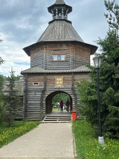 The image shows a wooden structure in Vzglyadnevo, Russia. It appears to be part of a historical site or park, with a bell tower and an arched entrance. The structure is made of stacked logs and has a weathered appearance. There's an icon above the archway. A paved pathway leads through the arch, and there are people walking through it. There is a red trash can nearby. A streetlight stands beside the structure. Green vegetation and trees surround the area. The sky is overcast.