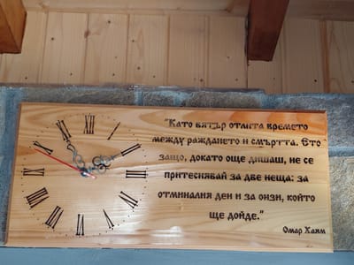 The image shows a wooden clock with a quote carved into it. The quote is in Bulgarian and appears to be attributed to Omar Khayyam. The clock has Roman numerals and a red second hand. The clock is mounted on a stone wall, with wooden paneling above it. The overall impression is of a rustic or traditional setting. It is not possible to determine the location in the city of Dryanovets, Bulgaria from the image alone.
