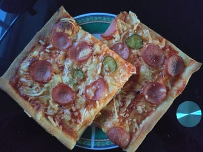 Two slices of pepperoni pizza are on a dark, reflective surface. The pizza slices are square-cut and overlap each other. Both slices are topped with tomato sauce, cheese, pepperoni slices, and pieces of pickle. The crust of the pizza is golden brown and appears to be thin. Underneath the pizza slices is a plate with a green and blue patterned rim. To the right of the pizza, there is a circular, metallic object that reflects a blue light. The background is dark and out of focus, suggesting the image was taken indoors, possibly in a kitchen or dining area. There are no people visible in the image. There is no information in the image to suggest the location in the city of Averøy, Norway.