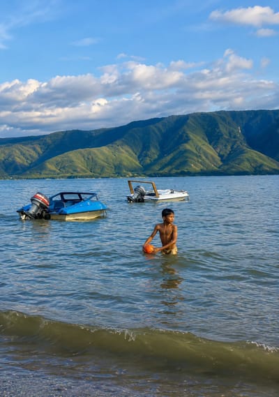 In Situngkir, Indonesia, a young boy stands waist-deep in the calm, blue waters of a lake. He holds an orange ball, looking towards the right. In the background, two motorboats are moored on the water: a blue boat with a red and black outboard motor to the left, and a white boat with a similar motor and a wooden frame to the right. Beyond the lake, a range of lush green mountains rises under a bright blue sky dotted with white, fluffy clouds. The sun casts shadows on the mountains, highlighting their contours. The water's surface has gentle ripples, and a small wave is breaking on the pebbled shore in the foreground.