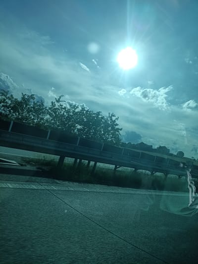 The image is a daytime outdoor photograph, captured from a moving vehicle, depicting a roadside scene in Třemošnice, Czechia. The upper two-thirds of the frame are dominated by a bright, partly cloudy sky, characterized by a distinct cyan-blue hue. A brilliant, circular sun, emitting a multi-point lens flare, is prominently positioned in the upper right quadrant. Scattered cumulus and wispy clouds are distributed across the sky, with a secondary, less intense circular light artifact or reflection visible in the upper left. A horizontal highway guardrail, featuring a dark metal rail atop vertical supports and backed by a solid barrier with intermittent white rectangular panels, spans the mid-ground. Behind the barrier, a dense line of mature deciduous trees forms a green border against the sky, indicating a raised embankment or hill. The lower third of the image presents a dark, textured road surface, likely asphalt, displaying horizontal streaking suggestive of motion, along with a subtle white lane marking. The immediate foreground on the right side contains a clear reflection of a vertically striped object, likely textile, appearing on the window from which the photograph was taken. No people are visible.