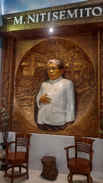 The image captures an indoor display within what is identified as the M. Nitisemito Museum complex in Glantengan, Kudus Regency, Central Java, Indonesia. Dominating the scene is a large, central, wall-mounted relief sculpture. Above this sculpture, a horizontal white sign with prominent capital letters reads "M. NITISEMITO."
The relief itself is predominantly brown-toned, with a central, three-dimensional bust of a light-skinned man. This man, presumed to be M. Nitisemito, wears spectacles and a traditional Indonesian blangkon (head covering). He is depicted in a white, long-sleeved, high-collared shirt with a small, dark emblem visible on the left side of his chest. His right hand is positioned across his upper abdomen. The background of the relief features intricately carved details of a traditional Javanese-style building surrounded by trees and landscape, all within a circular frame.
The wall behind the relief is a light, neutral color. Below the display, on a light-colored tile floor, two dark brown wooden chairs with slatted backs and armrests are positioned, facing the sculpture. Between these chairs rests an irregular, dark brown wooden object resembling a tree stump or a rustic stool. To the far left, a tall, dark vase with a decorative pattern contains several upright, slender, dry plant stems. A single overhead spotlight illuminates the main relief and signage.
