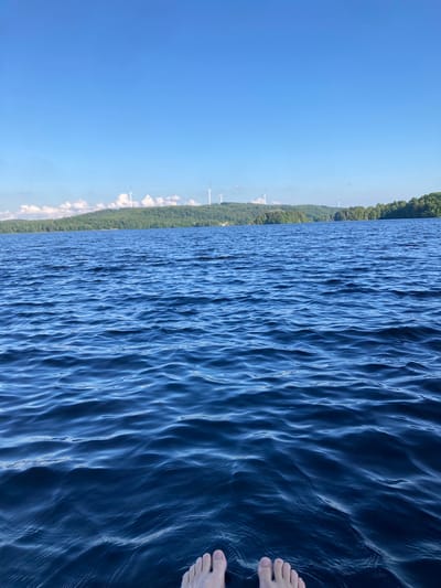 A pair of bare feet is positioned at the bottom of the frame, with the toes pointing towards the viewer. The feet are partially submerged in dark blue water that exhibits gentle ripples. The water fills the foreground and midground of the image. In the background, a green, tree-covered hill stretches across the horizon, dotted with several white wind turbines. The sky above is a clear, bright blue with a few scattered white clouds. The scene appears to be a natural landscape, likely a lake, in Vessigebro, Sweden.