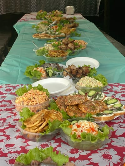 This image displays a spread of food laid out on a table covered with a light blue plastic tablecloth on the left and a red and white floral patterned tablecloth on the right. The food appears to be prepared for an outdoor gathering or picnic.

On the left side of the table, a series of clear plastic containers hold various dishes. Some contain what looks like fried chicken pieces and salads. The salads seem to be a mix of chopped vegetables like cabbage, carrots, and possibly cucumbers, dressed with a creamy sauce. Lettuce leaves are used as a garnish or base in many of the containers. There are also white paper plates stacked on the table.

The right side of the table features a larger spread. Prominently displayed is a sandwich or open-faced tart topped with slices of cucumber, dill, and what appears to be fish or meat. Next to this are several pieces of meatloaf or a similar savory dish, sliced and arranged. Further along, more clear plastic containers hold salads, similar to those on the left, one of which contains what looks like shredded carrots and a creamy dressing. Another container holds fried potato wedges, also garnished with lettuce.

The overall impression is one of a prepared meal or buffet, likely for a casual social event. The lighting suggests it is daytime. There are no visible people, and no text is discernible in the image. The setting appears to be outdoors, given the lighting and the nature of the food presentation. The location context is Надеждино, Russia.