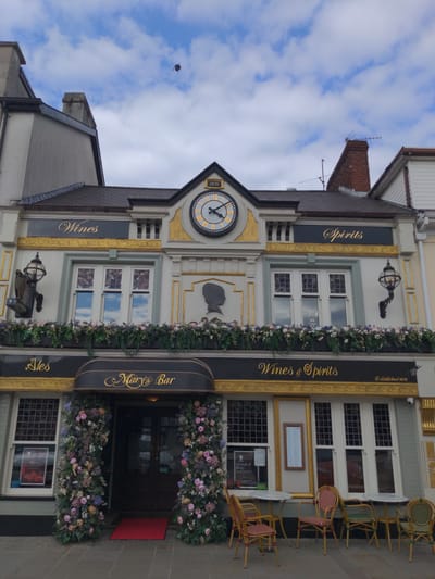 The exterior of "Mary's Bar" in Magherafelt, United Kingdom, is captured on a cloudy day. The building features a prominent clock with Roman numerals and a year "1876" above it. The facade is adorned with gold trim and signage for "Wines," "Spirits," and "Ales." The entrance to "Mary's Bar" is decorated with abundant floral arrangements. In front of the establishment, several outdoor tables and chairs are arranged, with a red doormat leading to the entrance. The street in front appears to be paved. The sky above is partly cloudy.