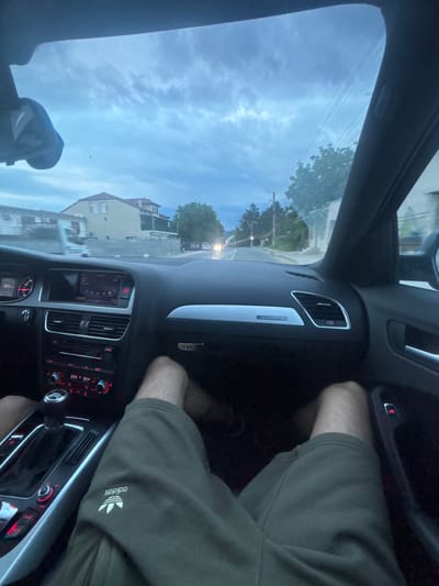 The image shows the interior of a car, specifically the driver's side dashboard and center console. The steering wheel, instrument panel, and climate controls are visible. The car appears to be an Audi, as indicated by the "Quattro" emblem on the dashboard. The driver is wearing green Adidas shorts. The car is moving in Daskotna, Bulgaria, as a street with buildings is seen through the windshield. The sky is cloudy.