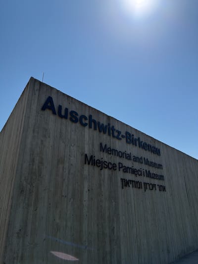 This media file shows the exterior of the Auschwitz-Birkenau Memorial and Museum in Oświęcim, Poland. The image is taken from a low angle, looking up at a stark concrete wall inscribed with text. The sky is a bright, clear blue, with the sun's glare visible in the upper left corner, suggesting it is daytime and likely a sunny day.
The concrete wall is textured, showing the imprints of the wooden planks used during its construction. Dark, raised letters are affixed to the wall, spelling out "Auschwitz-Birkenau" in English. Below this, there are lines of text in other languages: "Memorial and Museum", "Miejsce Pamięci i Muzeum" (Polish for "Place of Memory and Museum"), and Hebrew text.
The overall impression is one of solemnity and historical significance. The minimalist and imposing architecture, combined with the clear sky, creates a stark contrast that emphasizes the weight of the site's history. There are no people visible in the frame, focusing the attention entirely on the structure and its inscription. The image conveys a sense of presence at a place of profound historical importance.