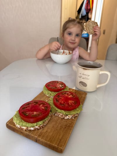 In Kavkazkaya, Russia, a young girl with blonde hair tied in pigtails sits at a white table, eating from a bowl with a spoon. She holds a crispbread in her other hand. In the foreground, three rice cakes topped with mashed avocado and tomato slices rest on a wooden cutting board. A mug with an image of a hedgehog and text that reads "in harmony with autumn" sits next to the cutting board. The scene appears to be a breakfast or snack setting.