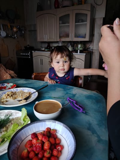 In a dimly lit kitchen, a young child sits at a turquoise table. The child, wearing a dark blue dress with cherry patterns and sporting a bandage on their forehead, gazes directly at the camera. In front of the child is a plate of food and a bowl of strawberries. A person's hand, with painted nails, is visible in the foreground, reaching towards the child. The kitchen environment shows cabinets, a stove, and various kitchenware, suggesting a domestic setting in the city of Блохино, Russia.