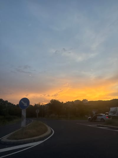 The image depicts a road intersection in Zagorsko, Bulgaria, during sunset. The sky is painted with vibrant hues of orange and yellow, transitioning to a soft blue at the top, indicating the approaching twilight. A round blue road sign with a white arrow pointing to the left is prominently displayed on the left side of the road. The road itself is asphalt, with white lane markings visible.
In the background, a cluster of houses is situated on a hillside, silhouetted against the colorful sky. Trees and vegetation line the landscape, adding a natural element to the scene. On the right side of the road, a silver car with its doors open is parked. Further down the road, a white truck is also visible. The overall impression is a serene, almost tranquil, roadside scene as day transitions into night in a Bulgarian setting.