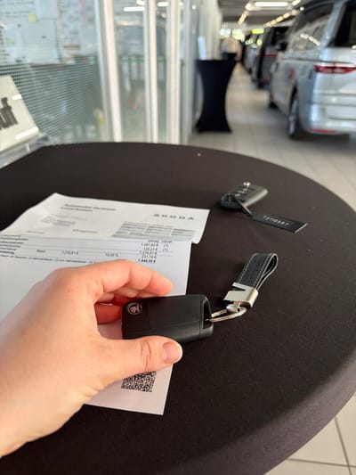 A person holds a Skoda car key and a document, presumably an invoice, in an automobile dealership. The document has the Skoda logo, references the city of Leverkusen, Germany, and itemizes costs, including a base price of 1,216.81 EUR, a VAT rate of 19.00%, and a final total of 1,448.00 EUR. The key has a black leather fob. Another key lies on a dark surface. Cars are visible in the background.