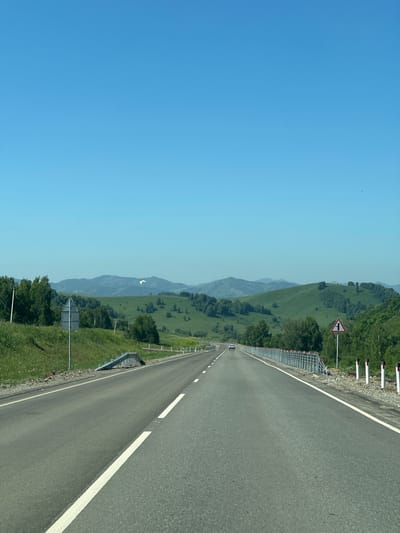 This image depicts a clear, sunny day on a paved road in Verkh-Aya, Russia. The road stretches into the distance, bordered by green, rolling hills covered in trees. A white double solid line marks the center of the road, with a white dashed line to the right. A guardrail with red and white reflective posts runs along the right side of the road. Several traffic signs are visible, including a triangular warning sign indicating a junction on the right. A white car is visible in the distance, traveling away from the camera. A single white bird is flying in the sky. The sky is a bright, clear blue.