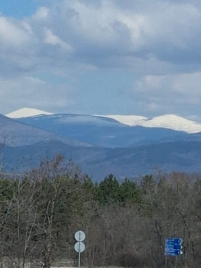 The image presents a landscape featuring a mountain range capped with snow on the left side, juxtaposed against a partially obscured view of what appears to be an urban edge on the right. On the right side, the presence of trees with bare branches, a road sign, and what might be buildings (though obscured) suggest this is a populated area rather than pure wilderness. The sky is visible and covered with clouds. Based on the query, the scene is in Ponor, Bulgaria.