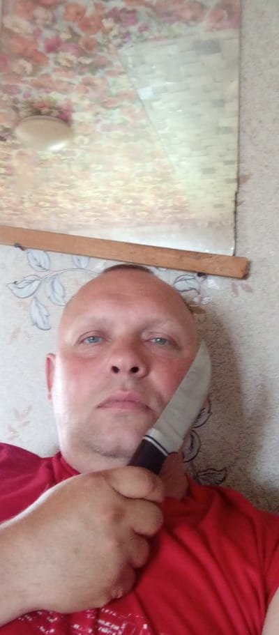 A close-up, low-angle, selfie-style shot shows a bald, fair-skinned man with blue eyes holding a knife against his left cheek. He is wearing a red t-shirt. The background is a wall with floral wallpaper and a wooden plank across the top. A reflective surface, possibly a mirror, is visible at the top of the image, showing a reflection of a patterned ceiling and a light fixture. The man's right hand, holding the knife, is visible in the foreground. The overall impression is that of an indoor setting, likely a residence.