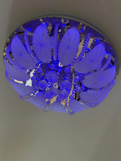 The image shows a modern, circular chandelier fixed to a neutral-colored ceiling. The chandelier features translucent, petal-shaped elements with radiating designs and strands of glass beads or crystals. Blue LED lights illuminate the structure. The location is unavailable from the visual data.