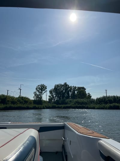 The image depicts a daytime scene from the stern of a white powerboat navigating a calm waterway. The sun is prominently visible as a bright orb in the upper-center sky, casting reflections on the water's surface. The sky is clear light blue with a few faint cirrus clouds and at least two visible airplane contrails. The opposite bank of the waterway is lined with dense green vegetation, including trees and bushes. Utility poles with overhead lines, consistent with power or railway infrastructure, are visible rising above the treeline on both the left and right sides of the background. The boat's interior, in the foreground, features white upholstered seating with dark blue accents and red piping on the left, and a brown, textured non-skid decking material on the gunwale to the right. Chrome grab handles are visible on the boat's sides. No individuals are visible. This scene is consistent with a view from the Sorraia River within the municipality of Coruche, Portugal.
