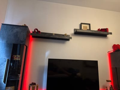 The image shows an interior space in Lüneburg, Germany, featuring a white wall with two floating shelves. On the left is a tall, dark cabinet with red LED lighting. A television is positioned centrally in the lower portion of the frame. The shelves hold decorative objects like a heart-shaped figurine, teddy bear figurines, a picture frame, and a box of roses. More decorative items are seen on top of the cabinet and on a small table to the left of the TV. The cabinet contains teddy bear figures behind a glass pane. Another cabinet is seen on the right with red LEDs and decorative items.