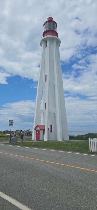 The image depicts the Pointe-au-Père Lighthouse, a prominent landmark in Rimouski, Canada, situated on the south shore of the St. Lawrence River. The lighthouse is a tall, white, octagonal structure with a red lantern room and a red-roofed auxiliary building at its base. Its design features vertical openings that create a lattice-like appearance. In the foreground, a grey asphalt road with a yellow center line is visible. To the left of the lighthouse, two individuals stand near a pedestrian crossing sign. The environment includes a grassy area, a rocky shoreline, and the expanse of the St. Lawrence River under a cloudy blue sky. The time of day appears to be daytime, given the lighting conditions.