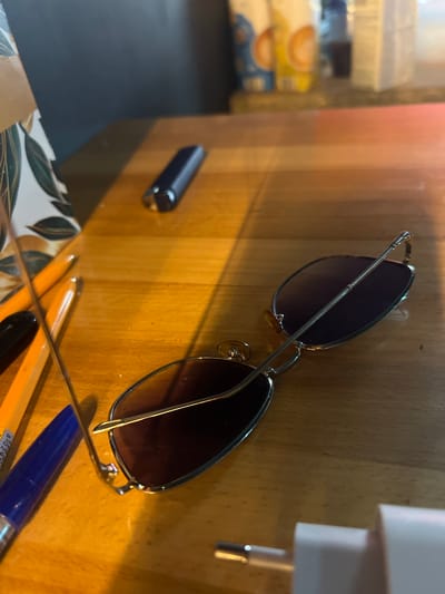 This is a close-up, eye-level shot of a pair of sunglasses with a thin metal frame and dark lenses, resting on a light brown wooden surface. The sunglasses are positioned diagonally, with the right lens lower than the left. To the left of the sunglasses, a black cylindrical object with a metallic end is visible, also resting on the wooden surface. In the foreground, parts of two pens are visible: one orange and one blue. A patterned surface, possibly paper or fabric with a leaf motif, is partially visible at the bottom left. In the background, out of focus, are stacked white items with orange circular designs, resembling packaging for food products. The lighting suggests a room with a warm light source, casting shadows on the wooden surface, indicating it could be daytime or evening. The overall impression is of personal items casually placed on a desk or table. There is no visible text in the image.