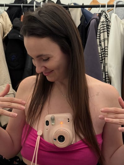 A woman is posing with a pink Instax Mini camera. She is wearing a pink top and a silver necklace. Her hair is long and brown. She is indoors with clothing hanging in the background. Her eyes are closed and she is holding her hand up in front of the camera.