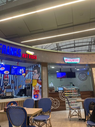 This is a photograph taken from within an indoor food court, likely at an airport or large commercial center, showing two fast-food establishments. On the left is "FRANKS HOT DOGS," featuring a blue and red neon sign and a graphic display of hot dogs and drinks. A staff member is visible behind a counter with a POS system. On the right is "Krispy Kreme DOUGHNUTS" with its distinctive green and pink logo. A display case filled with donuts is visible, along with a circular sign that reads "EAT 19 37 DOUGHNUTS." In the foreground, several tables and chairs with a blue and white patterned design are arranged for dining. The ceiling is illuminated by long, fluorescent light fixtures. The scene suggests a commercial area with food vendors and seating. The location is identified as Gollapalle Khurd, India.