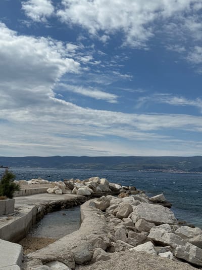 The image shows a rocky shoreline with a concrete pathway leading to the water's edge in Dugi Rat, Croatia. The sky is a clear blue with white clouds. The water is calm and blue, with a distant view of a green hillside across the bay. The rocky area surrounding the pathway is covered in white stones and pebbles. The pathway is slightly cracked and weathered.