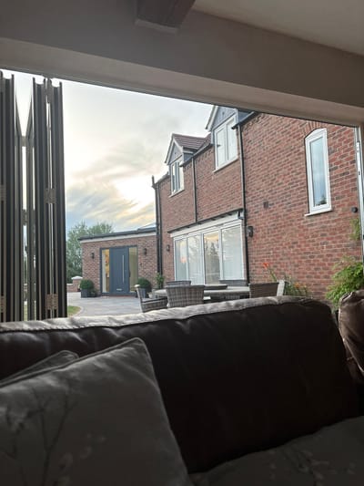 The image shows a view through an open folding door, looking out onto a neighboring house. The house is brick-built, with a modern grey door and a glazed extension. Outdoor furniture is visible on a paved area. The sky is light, suggesting it's either early morning or late evening. An out-of-focus brown leather couch with pillows is situated inside the room where the photo is taken.