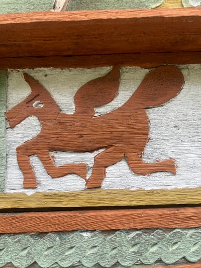 The image displays a close-up view of a wooden carving, likely a decorative element on a building facade. The carving depicts a stylized, winged horse or creature with a long, bushy tail, rendered in a rustic, folk art style. The carving is painted a reddish-brown color and is set against a white background, which appears to be a painted wooden surface. Above and below the carving are horizontal wooden planks, painted in various colors including dark red, yellow, and green. The green plank at the bottom features a decorative scalloped edge. The wood shows signs of age and weathering, with visible grain and peeling paint in some areas. There is a hint of a wooden beam above the carving. The setting is outdoors, suggested by the natural lighting and the weathered appearance of the materials. The image does not contain any individuals or indicate a specific action. The location in Meunasahmeucap, Indonesia is not visually evident from the image itself.
