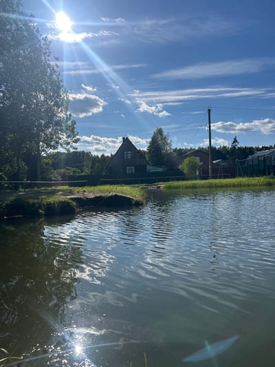The image depicts a scenic outdoor setting, likely in Слава Снт, Russia. The scene is dominated by a body of water, possibly a pond or small lake, reflecting the bright blue sky and scattered white clouds. Sunlight streams from the left side of the frame, creating a lens flare and illuminating the rippling surface of the water.
Along the shoreline, there is lush green grass and various trees, adding to the natural beauty of the environment. In the background, a traditional wooden house with a pitched roof is visible, suggesting a rural or semi-rural location. A power pole with wires is also present, indicating the presence of human infrastructure. The overall impression is one of a peaceful, sunny day in the countryside.
The time of day appears to be daytime, given the bright sunlight and clear sky. The weather seems to be fair and pleasant. There are no people or specific activities clearly depicted in the image, though the presence of the house implies habitation. The setting evokes a sense of tranquility and natural charm. No visible text is present in the image.