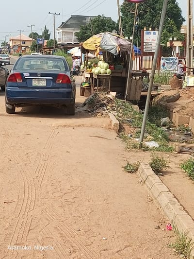 The image shows a blue Honda Civic parked on a dirt road in Ado Ekiti, Nigeria. The car is facing the camera. The road is lined with concrete curbs. There is a small stall to the right of the car with a variety of fruits, vegetables, and goods. Behind the stall is a tall metal pole with an object on top. The pole is leaning toward the road and is covered in vines. In the distance are buildings, trees, and a dirt road. The road is covered in tire tracks. There are several people standing and sitting in the distance, including one person standing near the stall. The image is taken at ground level, with the focus on the parked car. The coordinates (latitude: 7.659, longitude: 5.234) place the location in Ado Ekiti, Nigeria.
