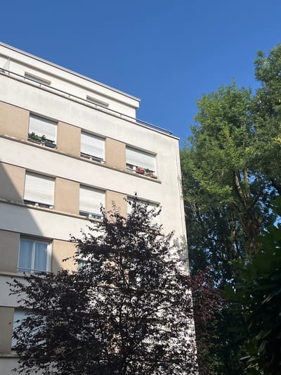 A medium shot shows a residential building in L'Haÿ-les-Roses, France, on a clear day. The building has a facade of beige and cream-colored panels, with multiple windows, many of which are covered by white rolling shutters. Some windows on the upper floors have planters with small green plants. A railing runs along the top of the building and balconies. To the right of the building are lush green trees, and in the foreground, a dark-leaved tree partially obscures the building. The sky above is a vivid, cloudless blue.