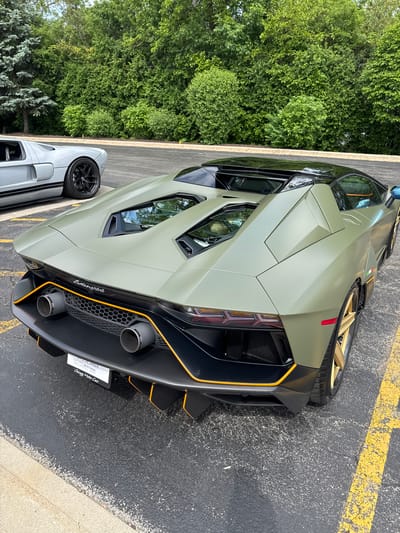 The image shows the rear of a matte olive green Lamborghini parked on an asphalt surface with yellow lines, likely in a parking lot. The vehicle has black accents with orange trim along the rear diffuser and around the exhaust pipes. A smaller, gray car is partially visible on the left side of the image, adjacent to the Lamborghini. In the background, dense green trees suggest a suburban or park-like setting. The license plate frame indicates "Chicago Motor Cars," and the license plate has an expiration date of "09/21/2026." Based on the license plate and dealership information, the location is likely in or near the city of West Chicago, United States, as Chicago Motor Cars has a location there.