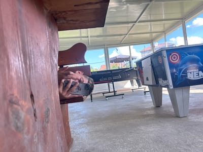 A young person is lying down on a wooden bench, holding a smartphone to take a selfie. The person is positioned near a wooden beam, with their face and hand visible. The background shows an outdoor covered area with a corrugated metal roof and glass walls. Through the glass, a bright blue sky with white clouds and some buildings and trees are visible. In the middle ground, two arcade games are present, one labeled "AJ-12" and another with "Finding Nemo" graphics. The floor is a light-colored concrete. The scene appears to be in a recreational area, possibly an arcade or a park, located in Glufishevo, Bulgaria.