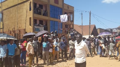 A crowd of people stands on a dirt road. Some hold umbrellas. Behind them is a three-story building with a balcony on the second floor. A metal frame structure stands in front of the building, supporting a sign. In the background, there are other buildings with brown roofs and a clear blue sky. The day seems sunny.
