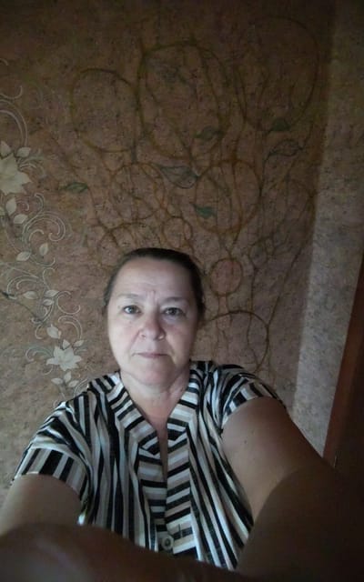 A medium-shot, slightly-angled, indoor, selfie-style photograph shows a woman with light brown hair pulled back and wearing a black, white, and tan striped blouse. She is positioned in the center of the frame, looking directly at the camera with a neutral expression. Her skin is fair and shows some signs of aging. The background consists of a textured wall with a muted brown and pinkish hue, featuring a subtle, darker, abstract swirling pattern. To the left of the woman, a decorative floral pattern with light beige flowers and swirling vines is visible. The lighting is somewhat dim and appears to originate from a source above and to the left, casting shadows on the right side of the scene. The overall impression is that of a casual, personal photograph taken in a domestic setting. There are no obvious clues to indicate the specific location within Zheleznogorsk-Ilimsky, Russia.