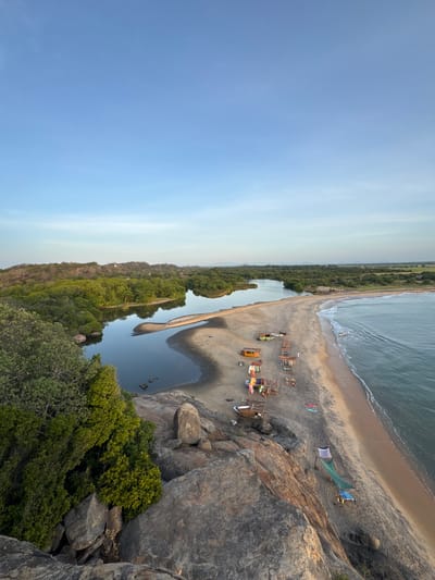 The image depicts a panoramic view of a coastal landscape in Shastrawela, Sri Lanka, at sunset. In the foreground, a rocky cliff offers a vantage point overlooking the scene. Below, a sandy beach stretches along the coastline, meeting the calm, blue waters of the Indian Ocean. A river mouth carves a distinctive shape into the beach, with lush green vegetation lining its banks and extending inland. Scattered along the beach are small, rustic shelters, some with surfboards and fishing equipment, suggesting a recreational or fishing settlement. The sky is a clear, soft blue, indicating the end of the day. A small boat is visible in the water, leaving a gentle wake. The overall impression is one of natural beauty and tranquility, with a hint of human activity integrated into the environment.