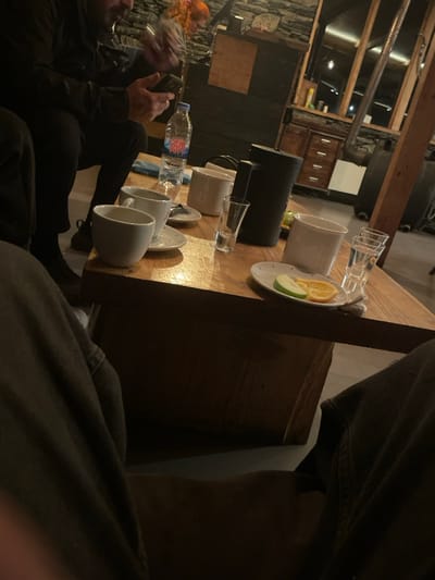 This image depicts an indoor scene within a rustic guesthouse or common area, consistent with the architectural style found in Juta, Georgia.

The foreground is occupied by the legs and dark trousers of a seated individual, presumably the photographer. Centrally positioned is a substantial rectangular wooden table, its surface reflecting ambient light. Upon the table are numerous objects: a plastic bottle of Rchveli brand water with a blue cap, two white ceramic cups resting on saucers, an additional white ceramic mug, a dark-colored insulated jug or French press, and two clear shot glasses, one appearing empty and another potentially stacked or containing a clear liquid. A small white plate holds a single slice of green apple and a single slice of orange. A blue fabric item is also visible on the table.

To the left of the table, a male subject is seated, visible from the chest up, dressed in dark clothing (jacket and trousers). He possesses a beard and is actively engaged with a smartphone, holding it with both hands and looking down at the screen. In the blurred background to the left, a third individual with reddish-orange hair is partially discernible.

The room's environment features a prominent wall constructed from natural stone blocks. Dark wooden cabinetry with multiple drawers is visible in the mid-background. Wooden structural beams are part of the ceiling or wall framework, and a metal pipe, possibly a stove flue, extends vertically. The lighting is low and warm, contributing to a cozy, somewhat dimly lit atmosphere.