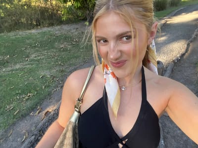 A medium-angle outdoor selfie shows a young blonde-haired woman in her late teens or early twenties, possibly of European descent, smiling with a slight nose wrinkle. She has fair skin, light eye makeup, and light pink lipstick. She's wearing a low-cut black halter top, a gold choker necklace with a clear pendant, and a patterned scarf tied around her neck. A beige designer handbag with a gold strap is slung over her left shoulder. The background includes a grassy area, trees, and a paved path. The photo was taken in daylight in an outdoor environment that might be Livingstone, Zambia.