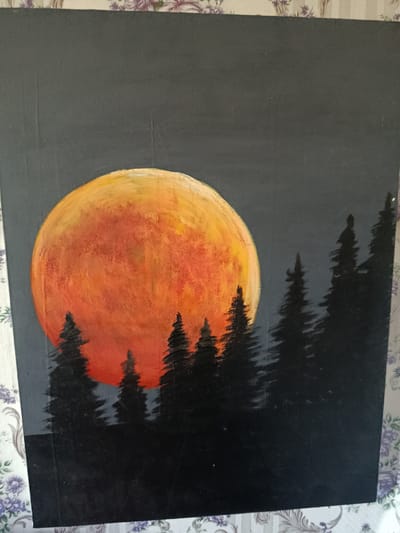 This is a photograph of an acrylic painting on a canvas. The painting depicts a surreal night scene with a large, vibrant orange and yellow moon dominating the left side of the canvas. The moon is textured, with visible brushstrokes giving it a luminous quality. Silhouetted against the moon and a dark gray sky are a row of pine trees. These trees are painted in shades of black and dark gray, with pointed tops characteristic of evergreen trees. The foreground is also painted in dark shades, suggesting a forest floor or the base of the trees. The overall impression is one of a dramatic and slightly eerie natural landscape under a large, intense moon. The painting is framed by a floral patterned cloth on the top and bottom. No text is visible in the painting or the photograph.
