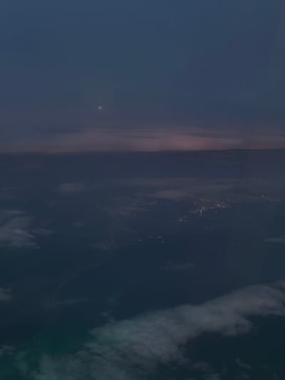 An aerial photograph depicts a nighttime or late twilight scene over an urban area within the Cotswold District, United Kingdom. The upper portion of the image shows a dark blue to deep purple sky, featuring a prominent, nearly full moon in the upper-middle. A horizontal band of lighter, diffuse clouds or atmospheric haze spans the mid-ground, with a faint pinkish glow visible above it, suggesting reflected light. Below this band, extensive patterns of bright, yellowish-white artificial lights illuminate a city or town's infrastructure, including roads and aggregated structures, set against the dark, indistinct terrain. Patches of white, wispy clouds partially obscure the lower foreground. No individuals or specific actions are discernible within the frame.